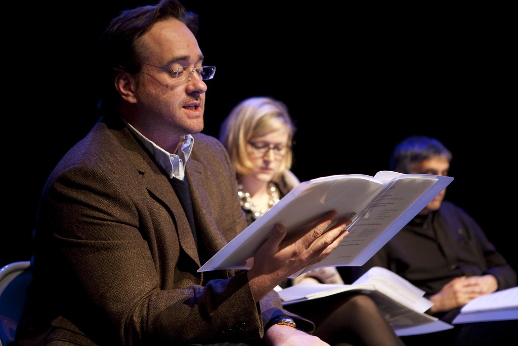  Matthew Macfadyen, dressed in a brown suit and wearing glasses, reads from a folder he holds in his left hand.