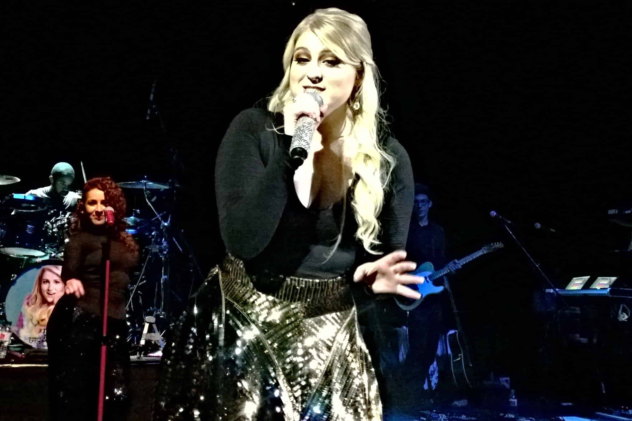 Meghan Trainor in a black top and golden skirt, holding a mic in her hand and singing at her concert