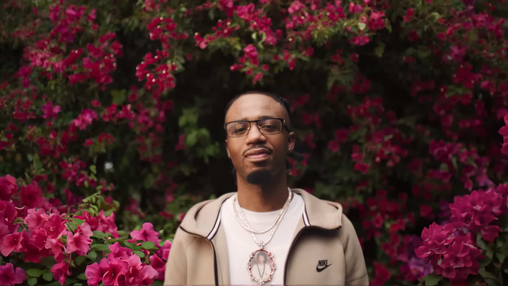 Metro Boomin wearing a Nike light brown jacket, standing in front of pink bougainvillea trees 