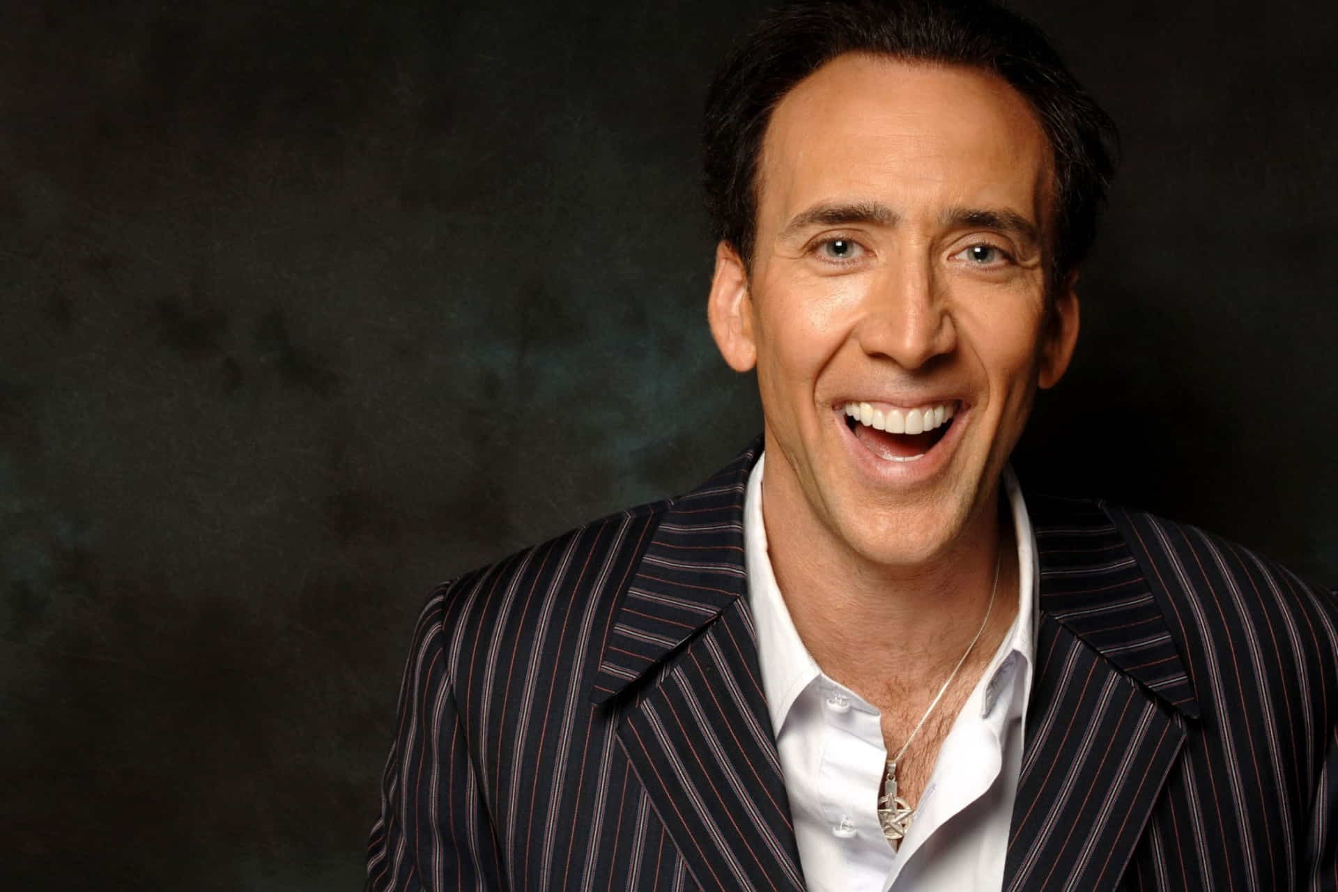 Nicolas Cage is wearing a striped suit with a white shirt and smiling at the camera.