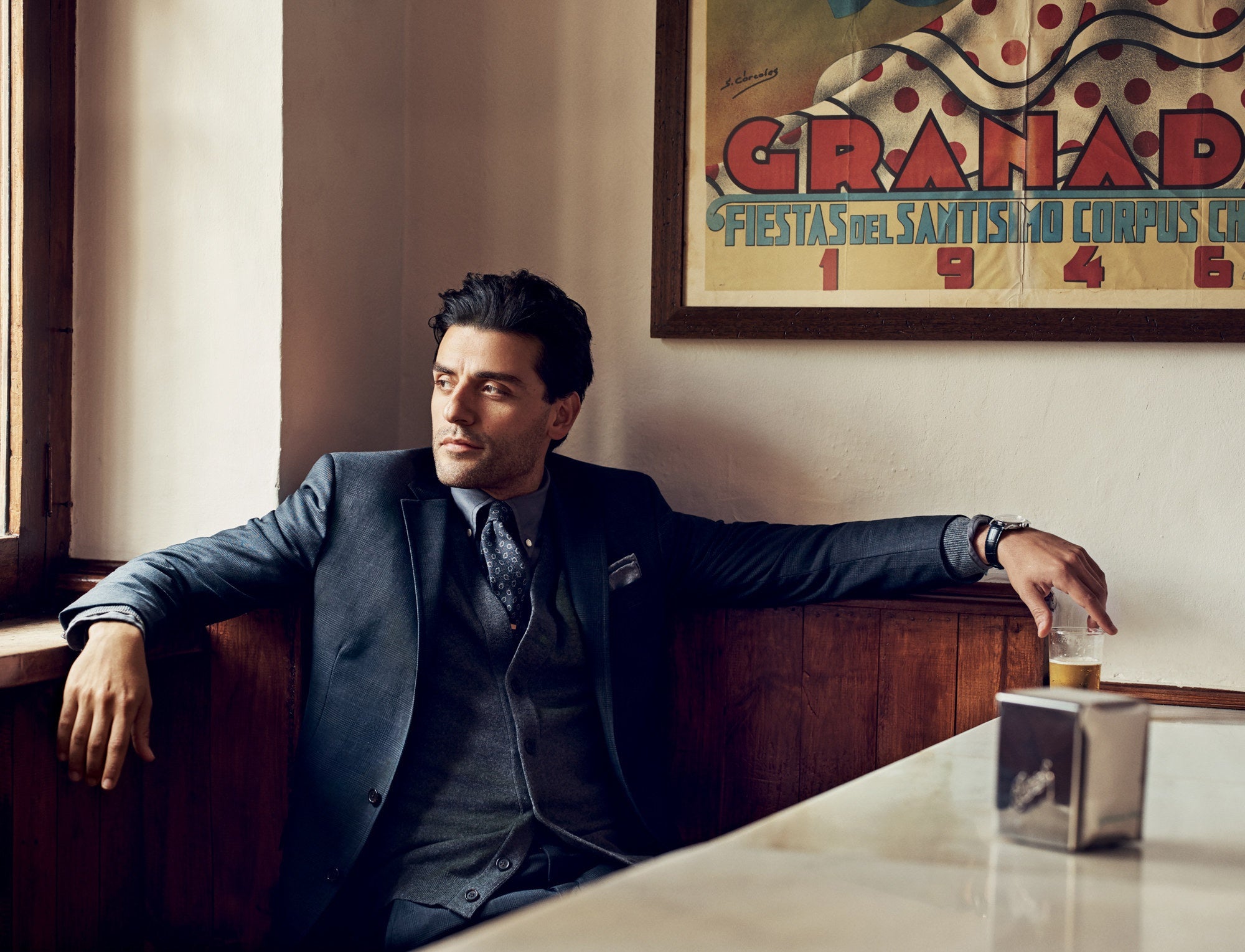 Oscar Isaac, wearing a navy blue suit, looking out the window with a glass of drink in hand