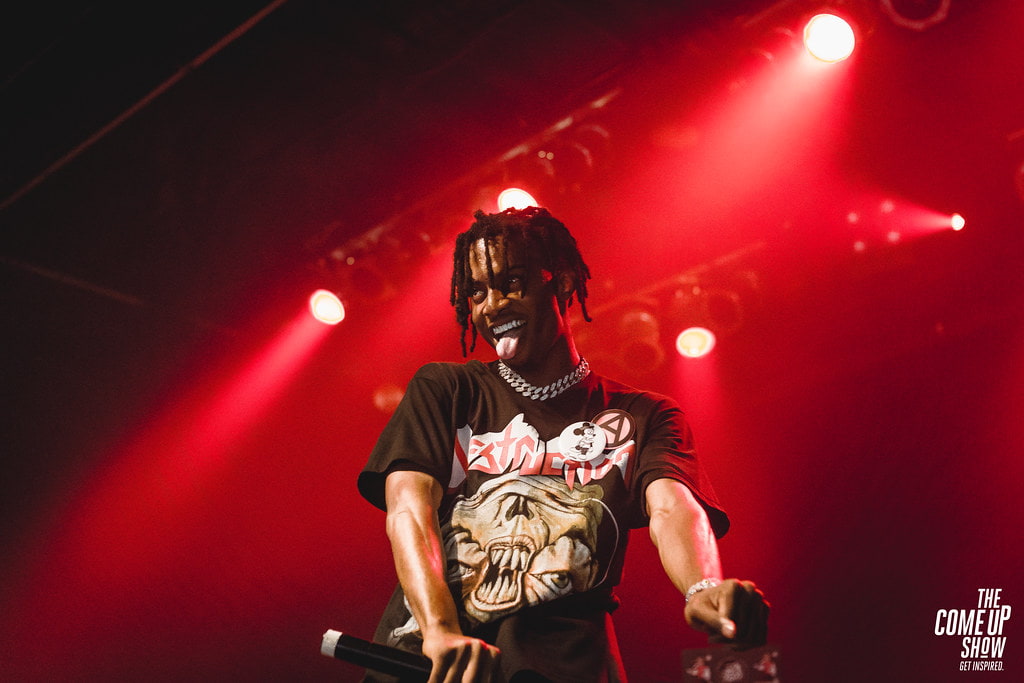 A high-contrast photo of Playboi Carti performing on stage, leaning forward with a microphone in hand.