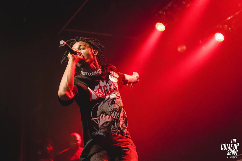 Playboi Carti is singing at a concert holding a microphone and there's a red background behind him.