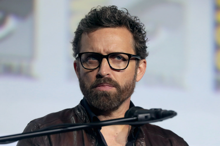 Rob Benedict, with a very intense expression, listening to the speaker at a press conference.