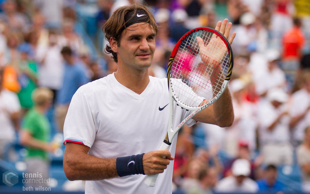 Roger Federer, wearing a white shirt and black headband, holding his tennis racket in his right hand and waving to the crowd with his left hand.