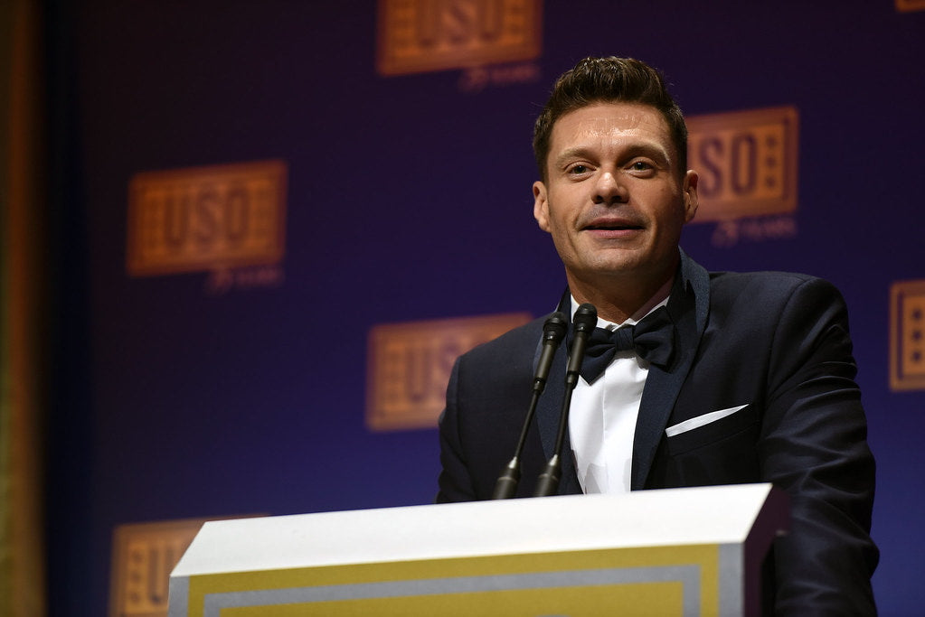 Ryan Seacrest in a black tuxedo, standing in front of a podium, and addressing the crowd