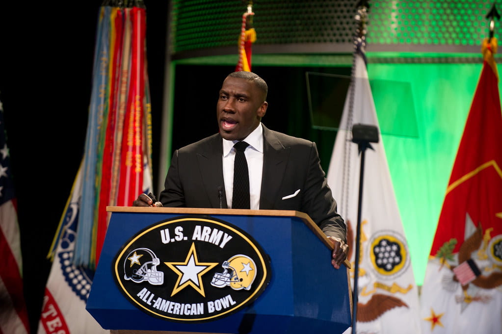 Shannon Sharpe in a black suit and tie standing at a podium with a U.S. Army All-American Bowl logo on it.