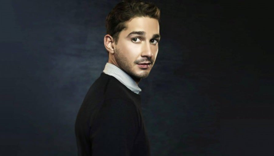 Shia Labeouf in a black sweater, posing in front of a  dark navy blur background