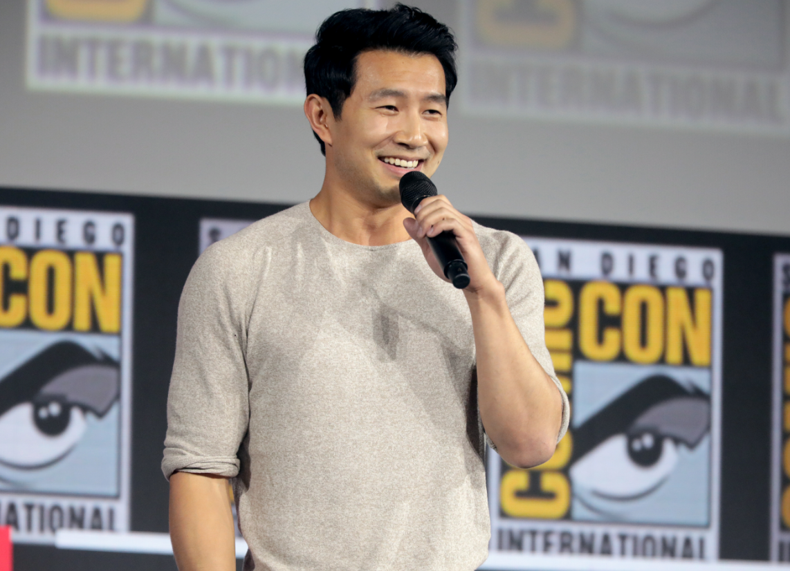 Simu Liu at a comicon holding a mic and addressing the crowd.