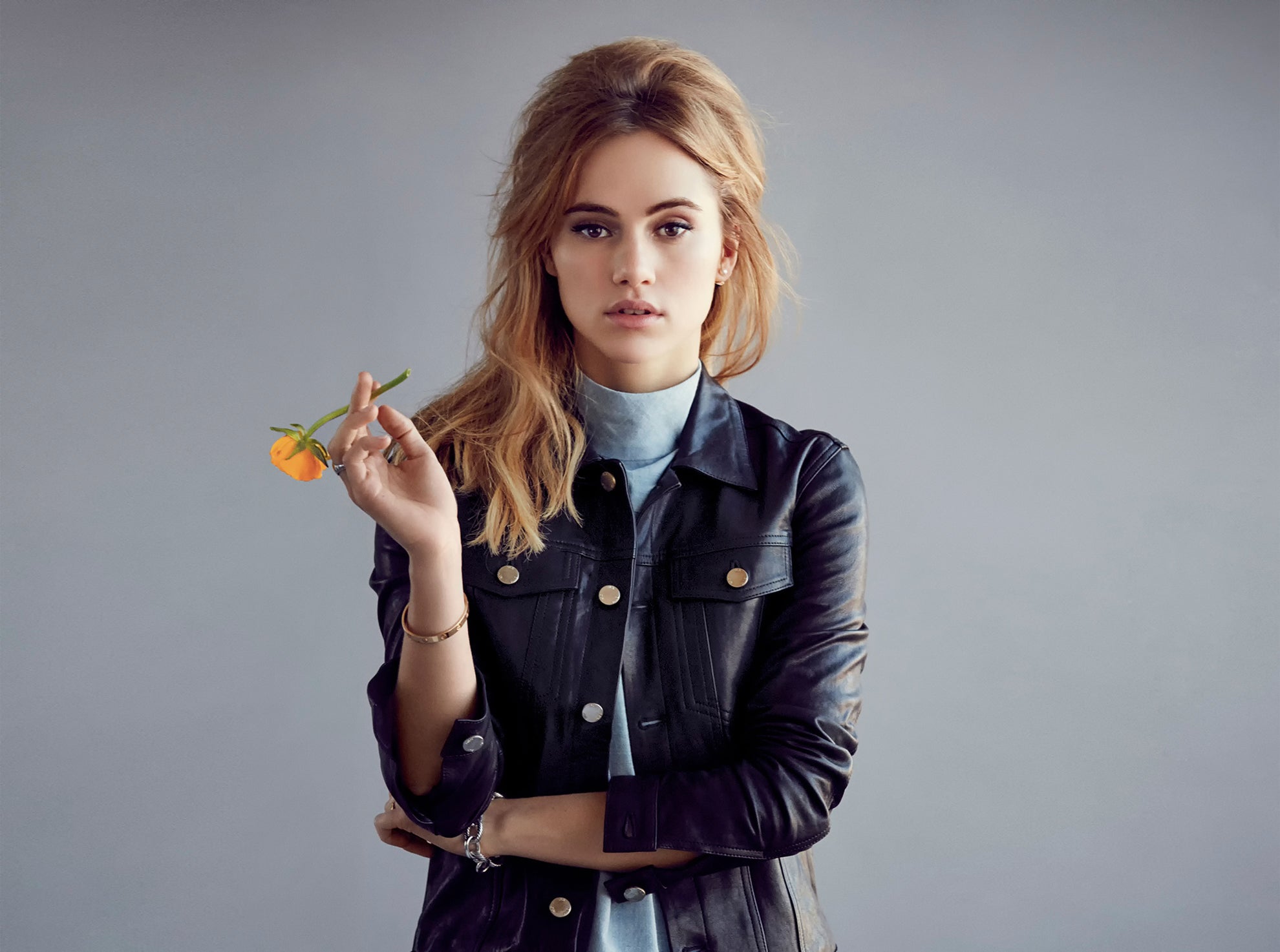 Suki Waterhouse holding a flower. She is standing indoors, wearing fashionable clothing and is the focus of a photo shoot.