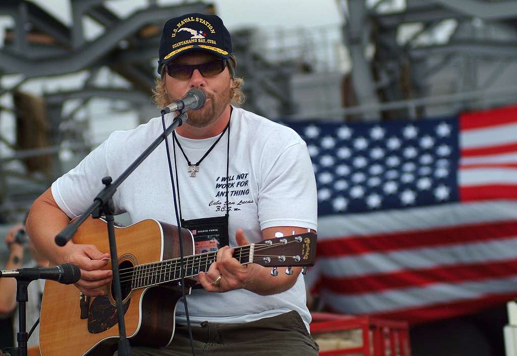 Toby Keith holding a guitar in his hand, wearing a white t-shirt and black shades, and singing at his concert with America's national flag in the background.