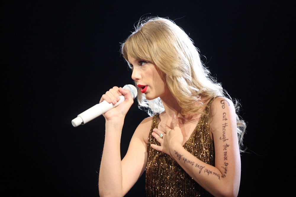 Taylor Swift in a gold shimmer dress, holding mic in her hand and singing at her concert.