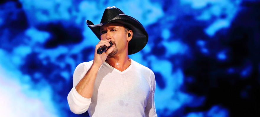 Tim Mcgraw in a cowboy hat, holding a mic in his right hand and singing at his concert