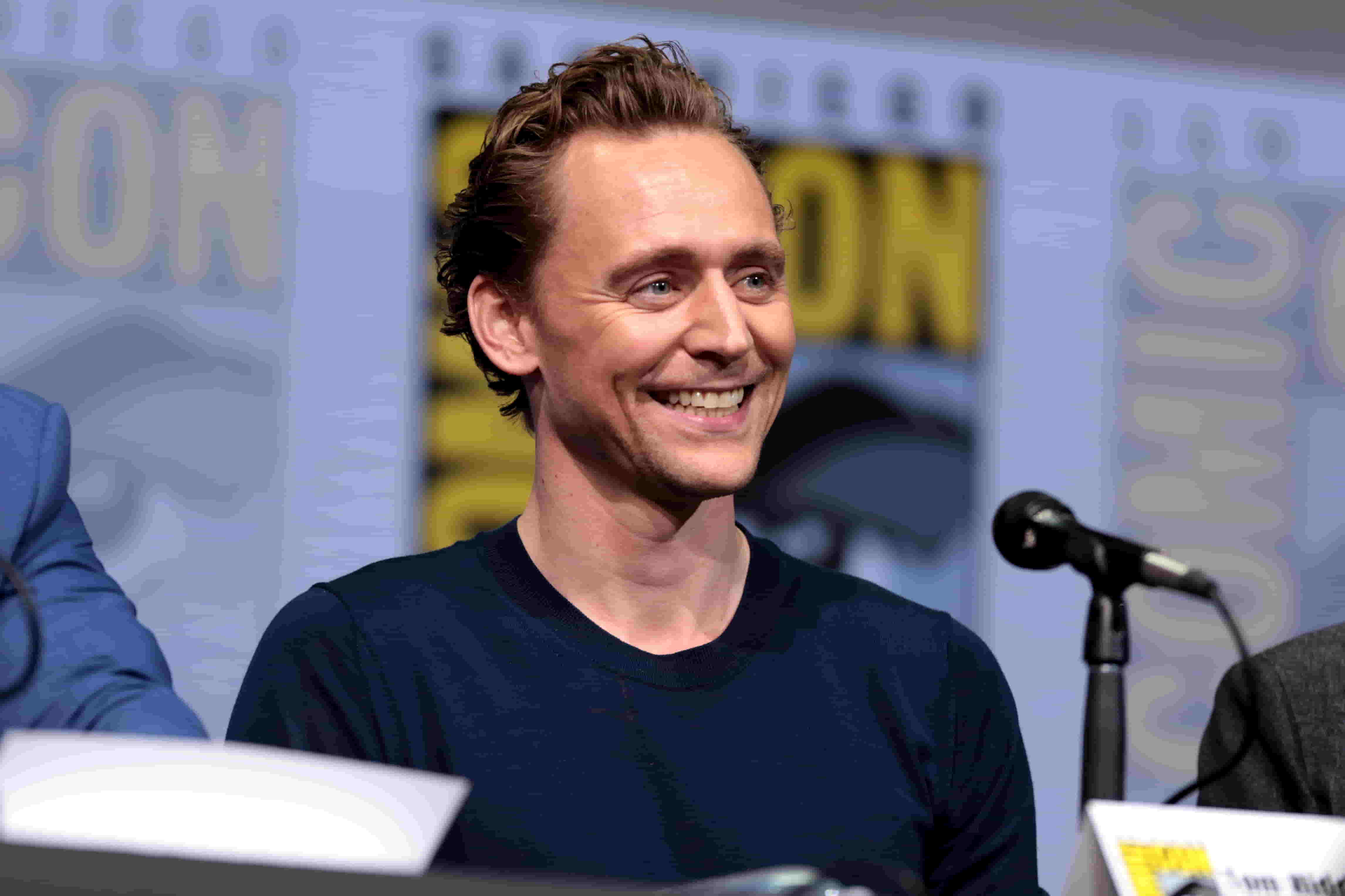 Tom Hiddleston is in a dark blue t-shirt at a Comicon event, sitting with a microphone kept in front of him