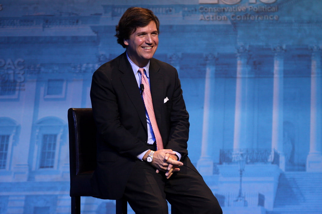 Tucker Carlson in a black suit, smiling while addressing the crowd