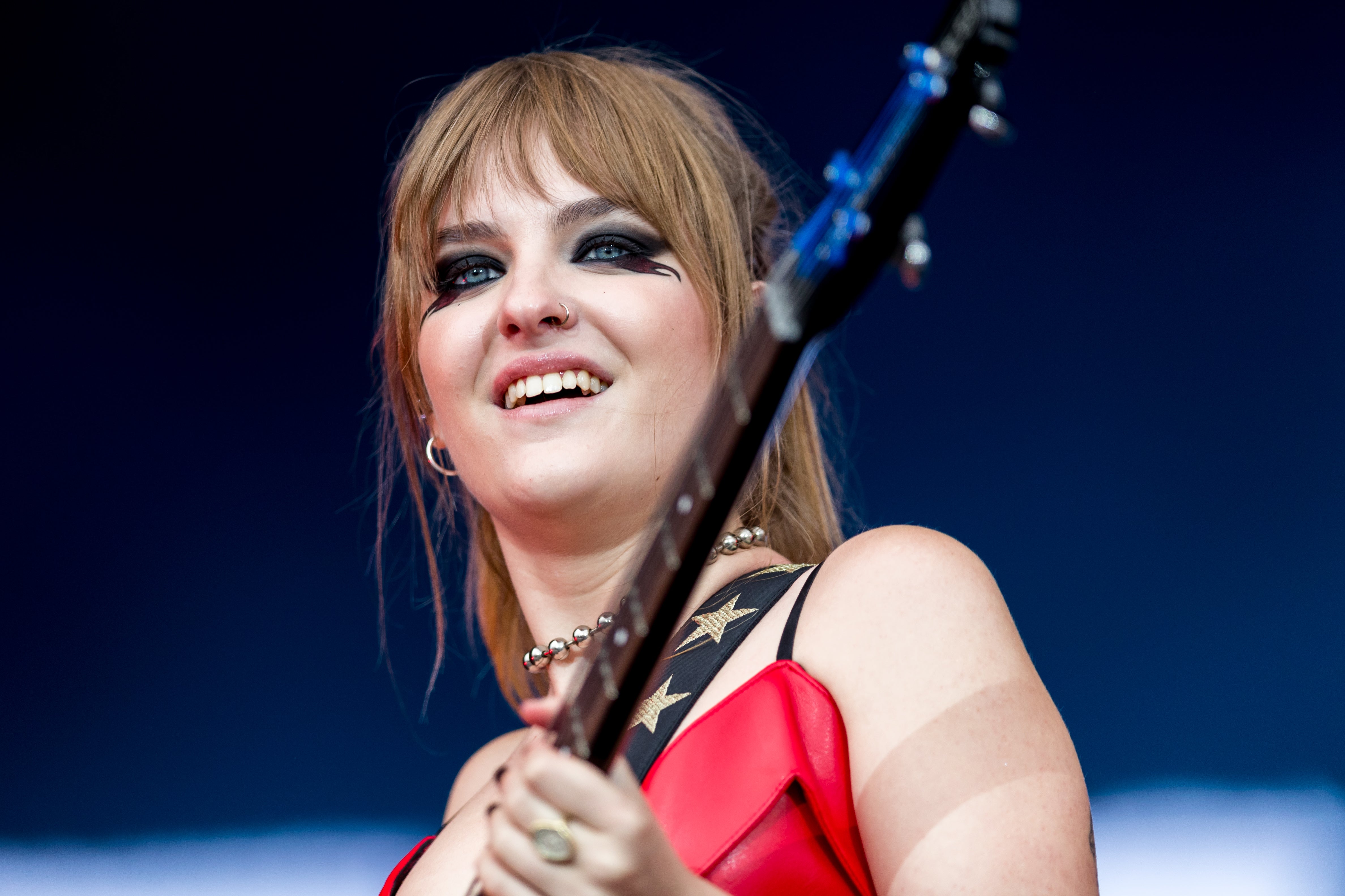 Victoria De Angelis smiling and playing an electric guitar on stage with a live audience.