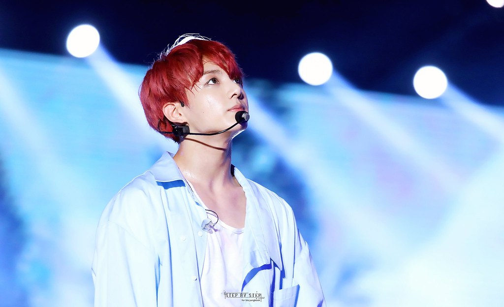 Jungkook is at the BTS concert, wearing a white shirt, looking upwards while performing on stage.