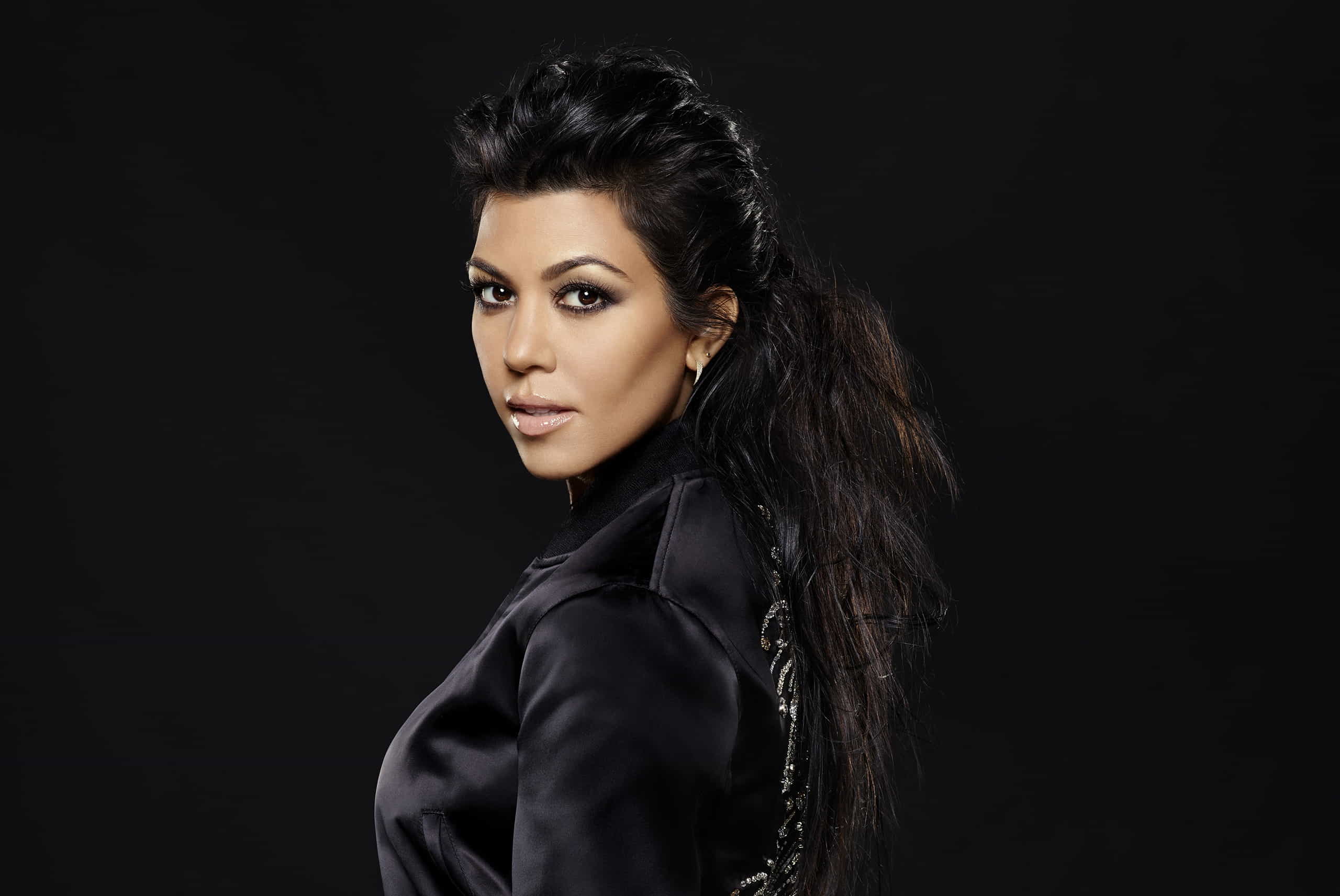 Kourtney Kardashian wearing a black dress standing in front of a black background and posing for a photo shoot.