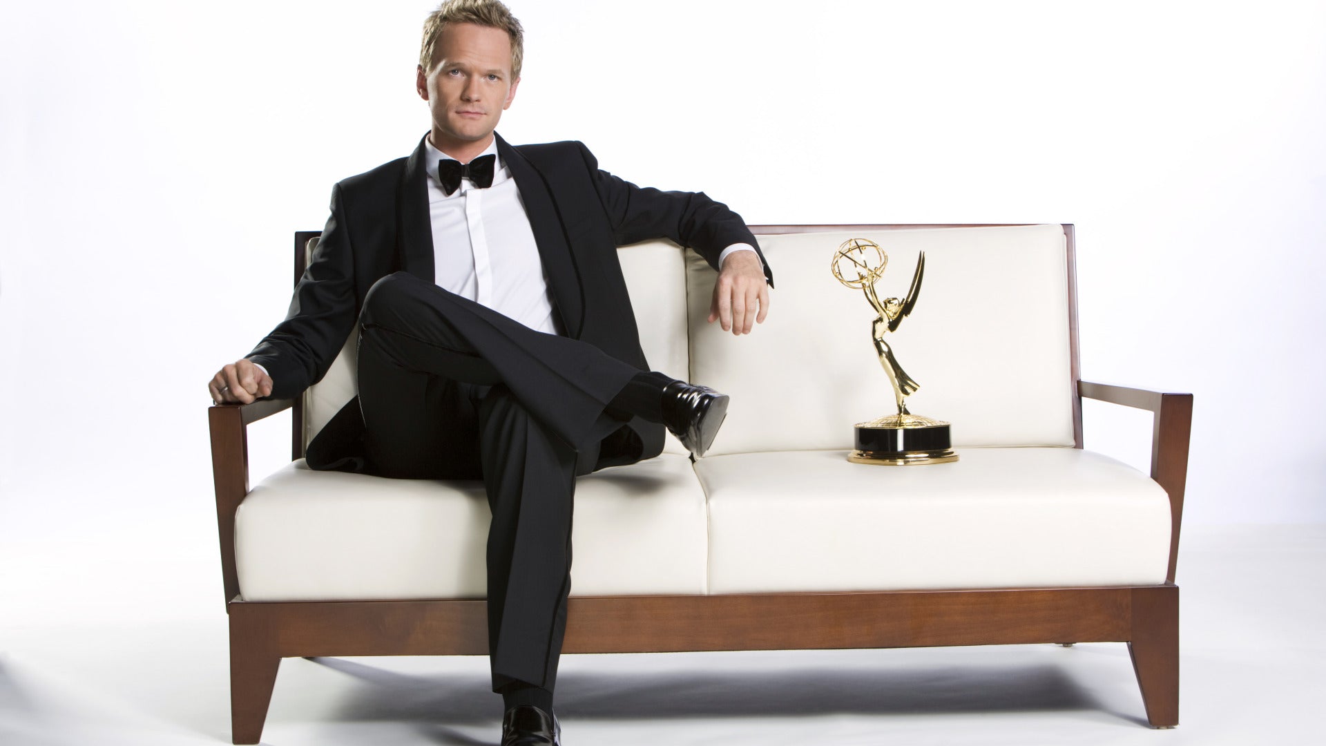 Neil Patrick Harris wearing a formal attire and sitting on a white couch.